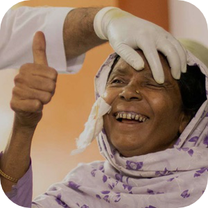 一位头戴碎花头巾的老年妇女喜笑颜开，医护人员戴着手套为她轻轻检查眼睛。她竖起大拇指，表达幸福和感激之情。她的围巾里塞着一张纱布，表明她最近接受了一次医疗手术。